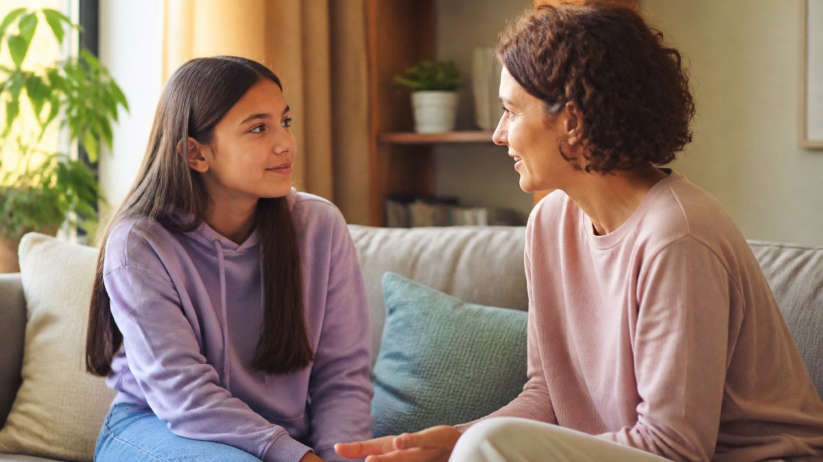 A parent and teenager having a calm conversation about adolescent mental health on a couch at home.