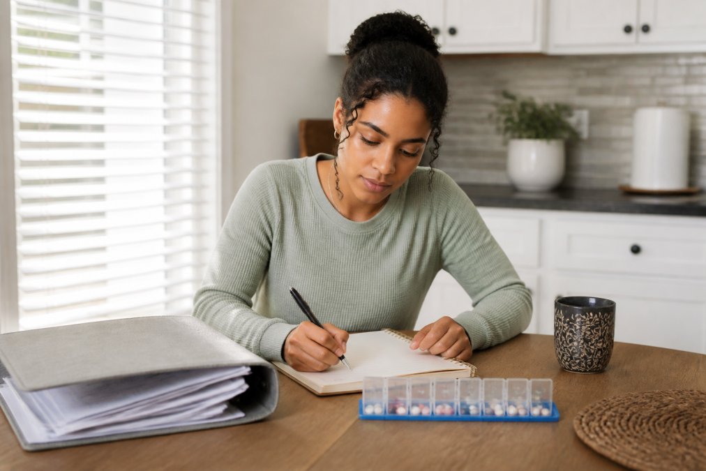 A person sitting at a kitchen table with a folder of medical records, a pill organizer, and a notebook, writing a list in a calm, bright room