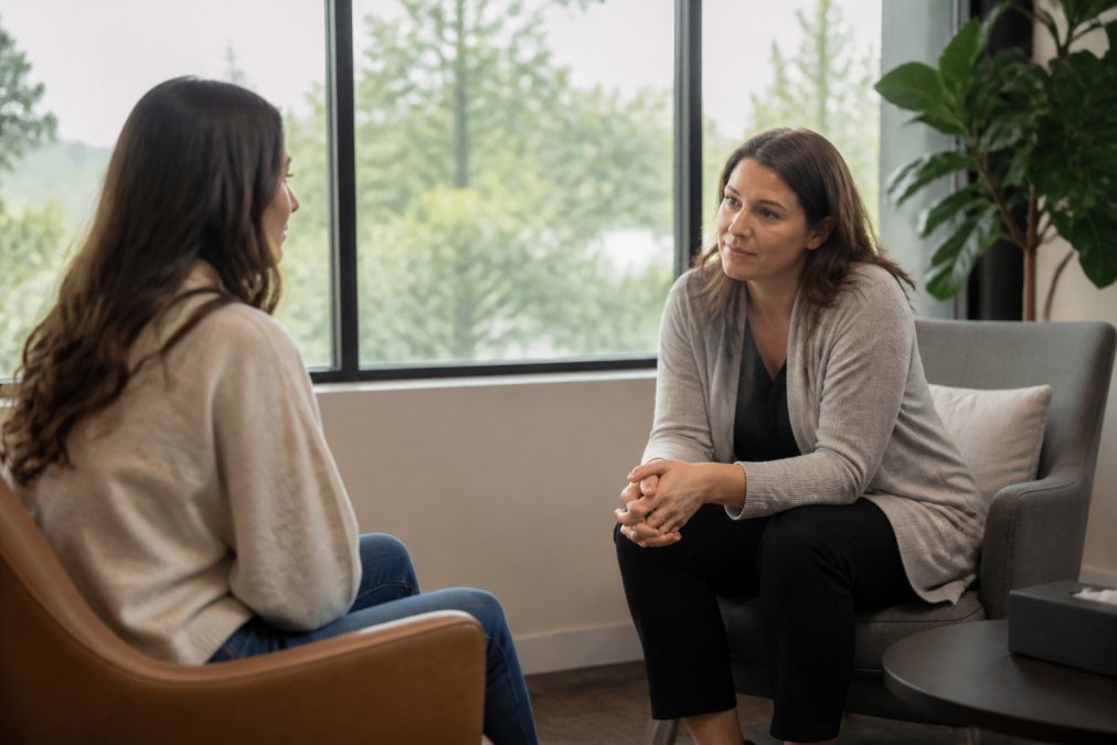 A warm, welcoming psychiatric office with a compassionate clinician leaning forward attentively while listening to a patient seated across from them in comfortable chairs, soft natural light coming through large windows behind them